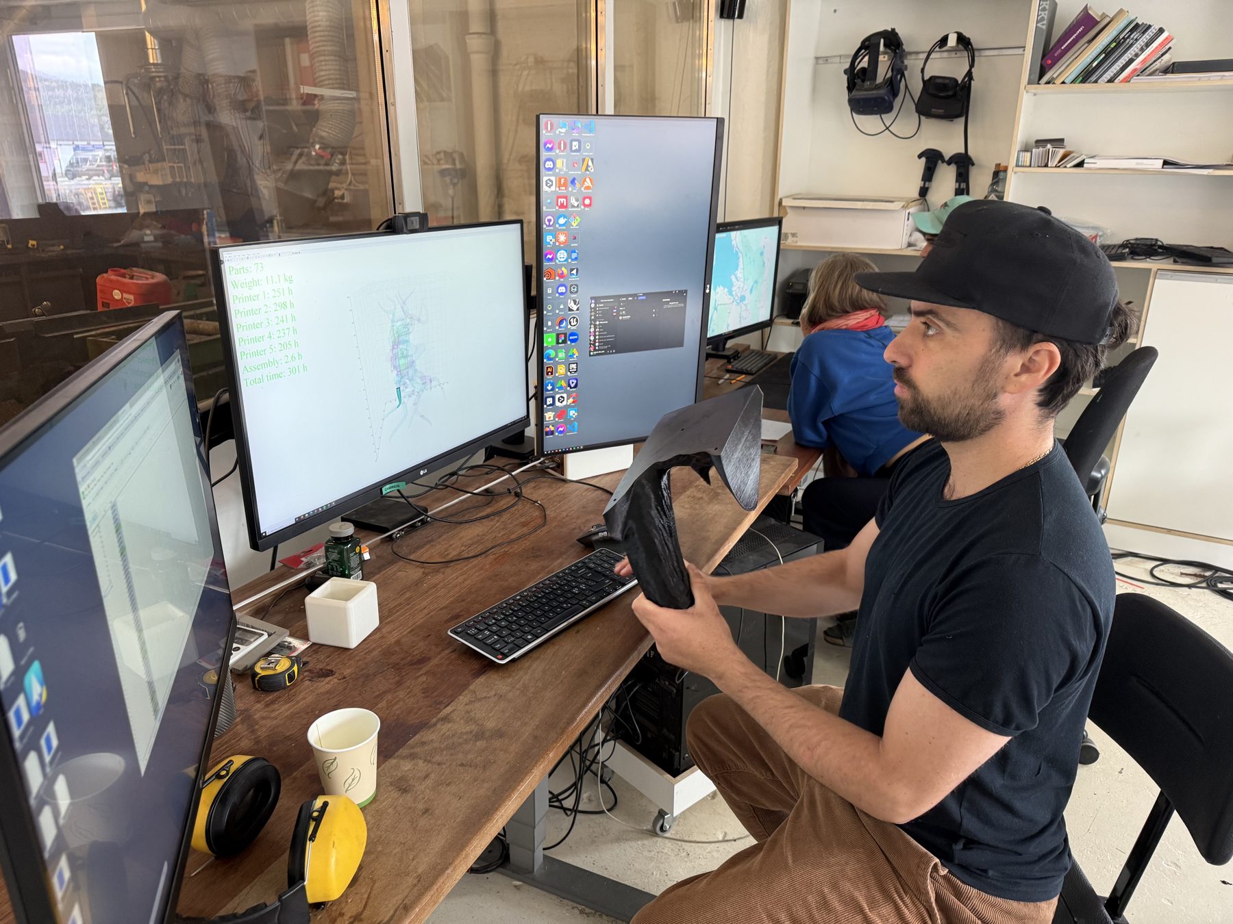 Someone at a bank of monitors holding a printed part, VR headsets on the wall behind