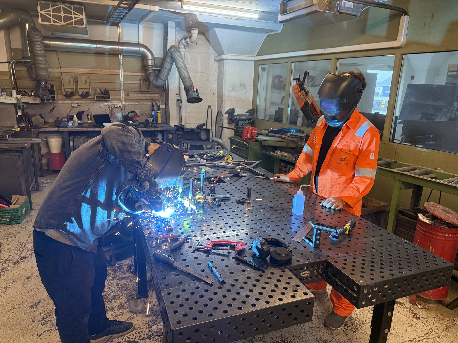 Two people welding at a steel fixture table in the metal workshop