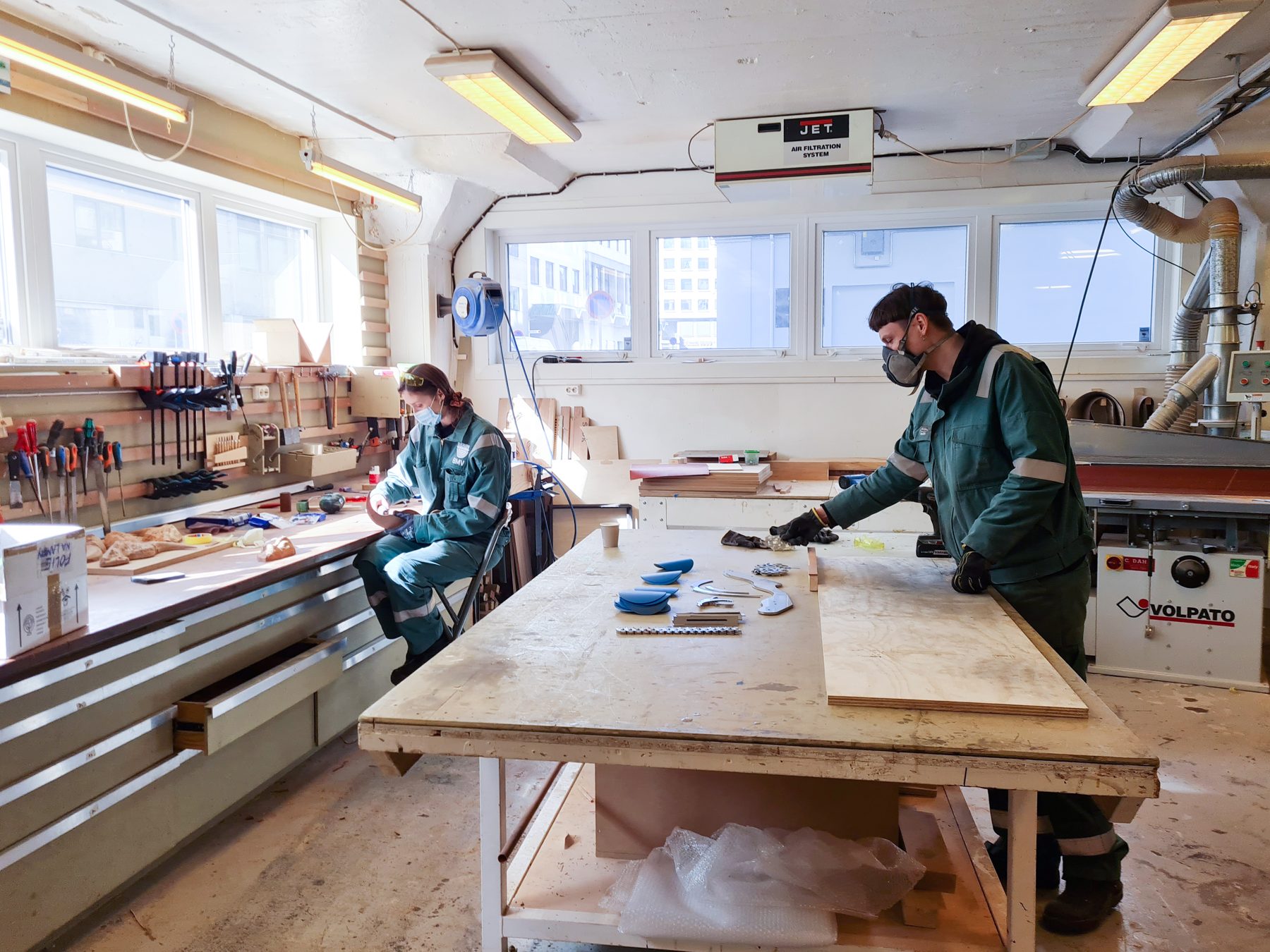 Two people working at benches in the wood workshop, harbour light through the windows