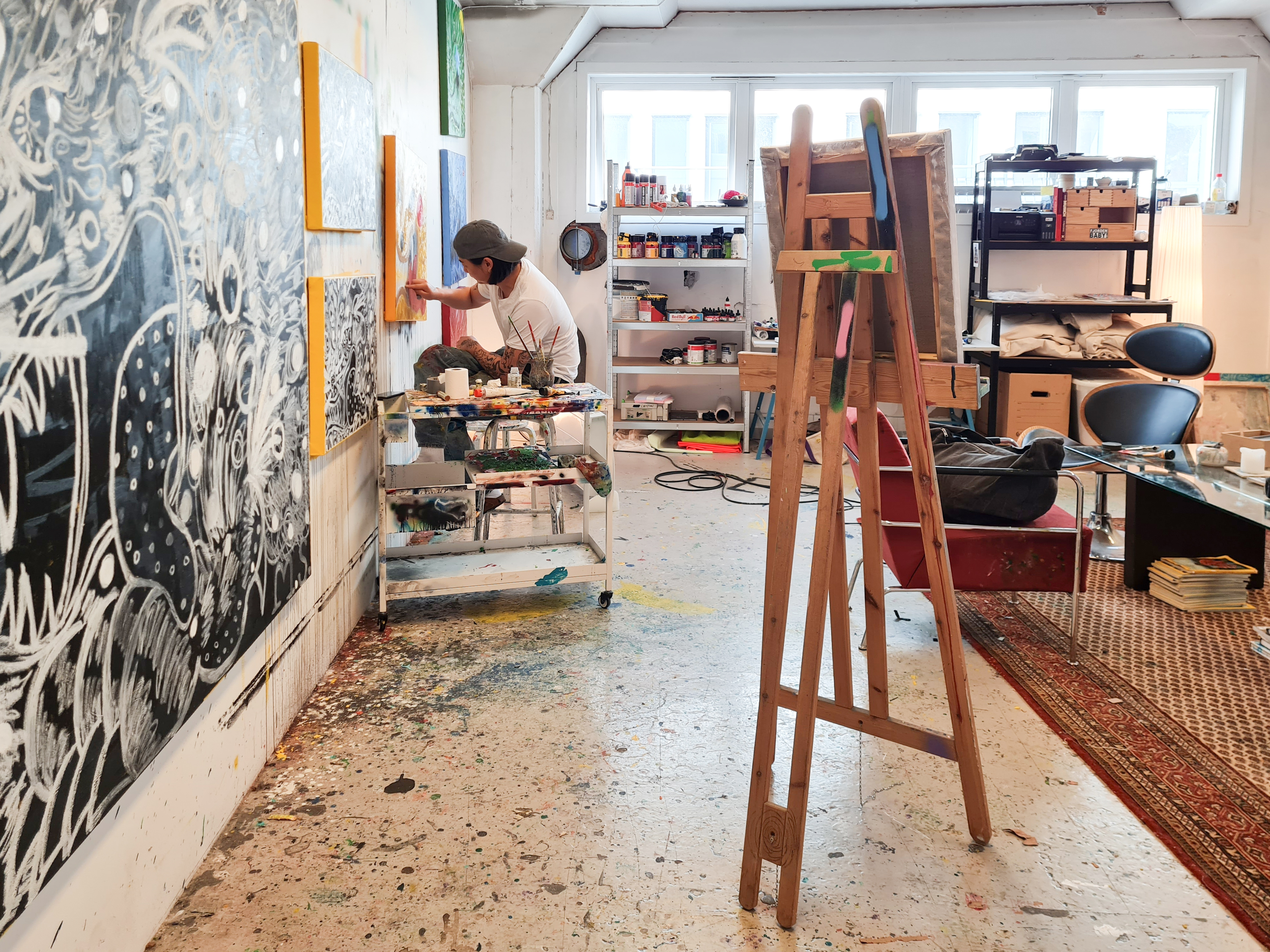 An artist at work in a shared studio at C. Sundts gate 55, painting against a wall of canvases, with a wooden easel and worktables across the room.